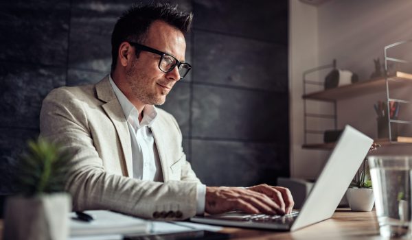 Businessman sitting at his desk and using laptop in the office Businessman wearing linen suit and eyeglasses sitting at his desk by the window and using laptop in the office
