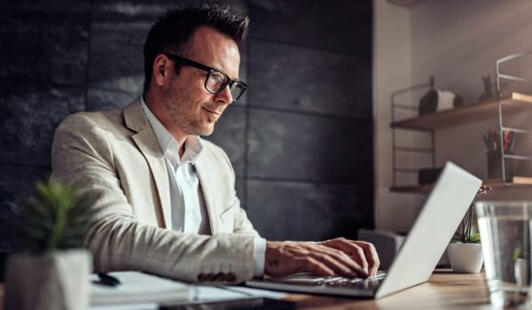 Businessman wearing linen suit and eyeglasses sitting at his desk by the window and using laptop in the office